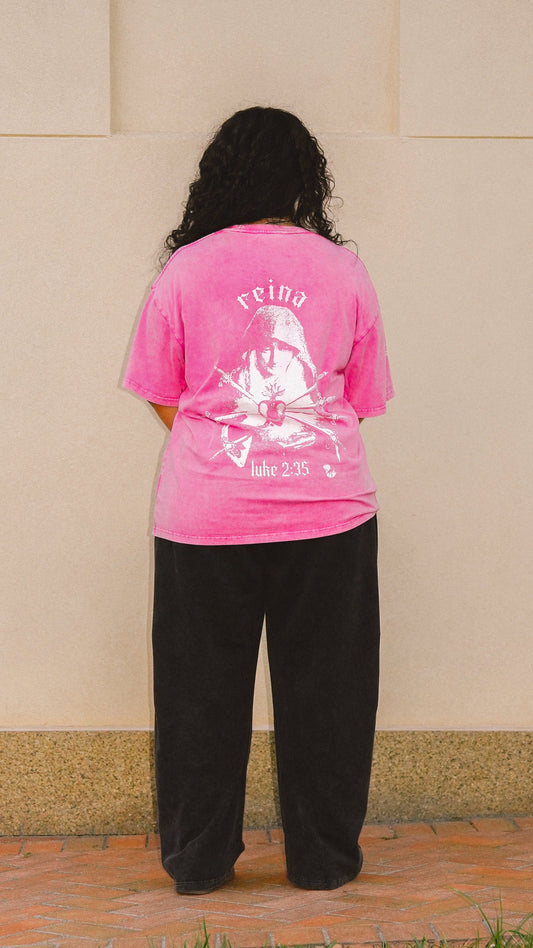 A person viewed from behind wearing a bright pink graphic t-shirt with an image of the 7 sorrows of Mary with the word Reina above her head and loose black pants, standing outdoors against a beige wall.