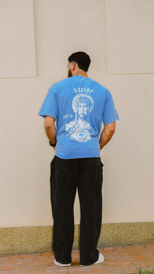 A man viewed from behind wearing a light blue graphic t-shirt with a large white illustration of Christ and the word 'Savior' on the back, paired with black pants and white sneakers, standing against a beige wall.