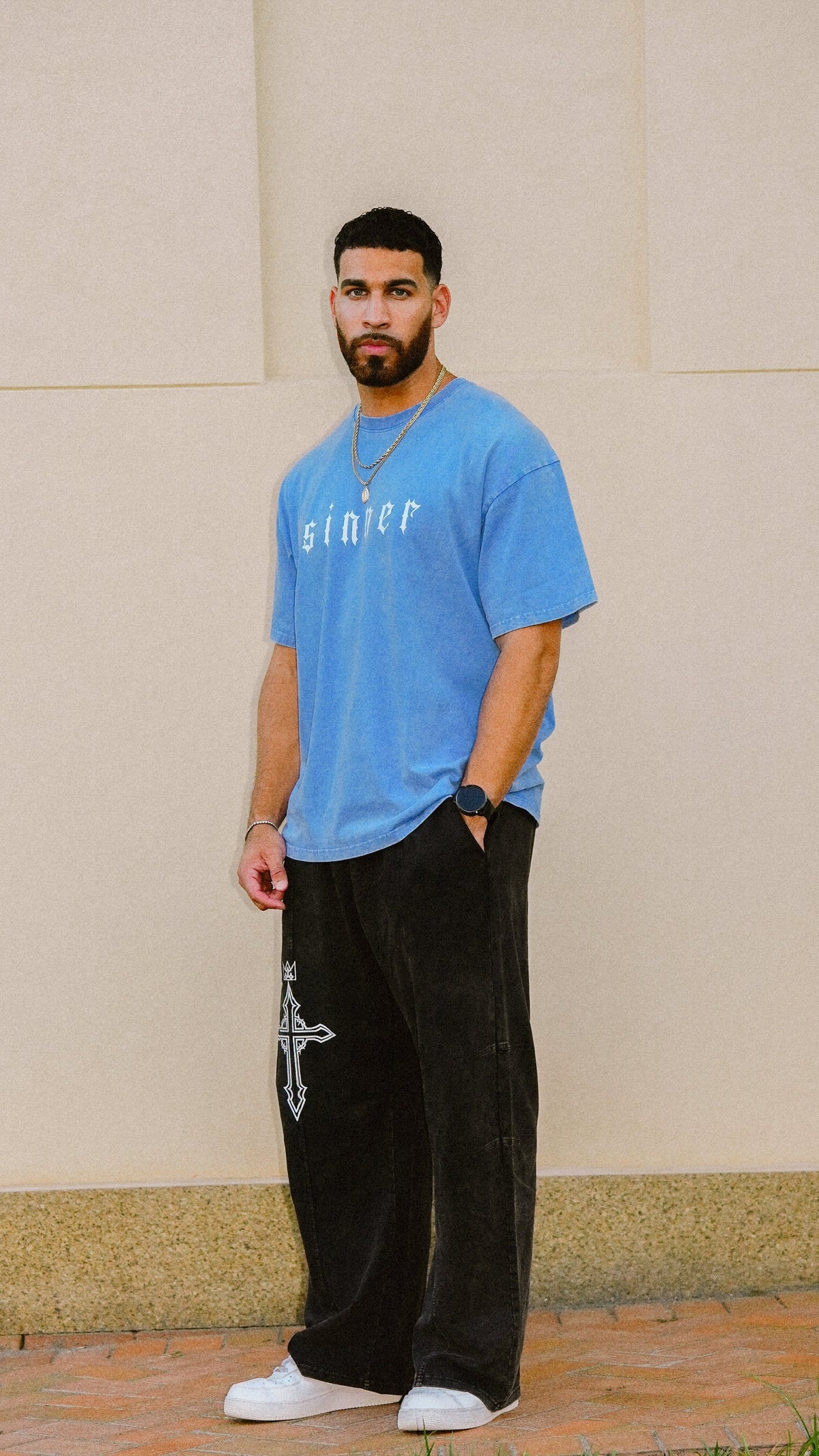 A man wearing a light blue t-shirt with 'sinner' printed on the chest, black wide-leg pants with a white cross graphic, and white sneakers, standing against a beige wall.