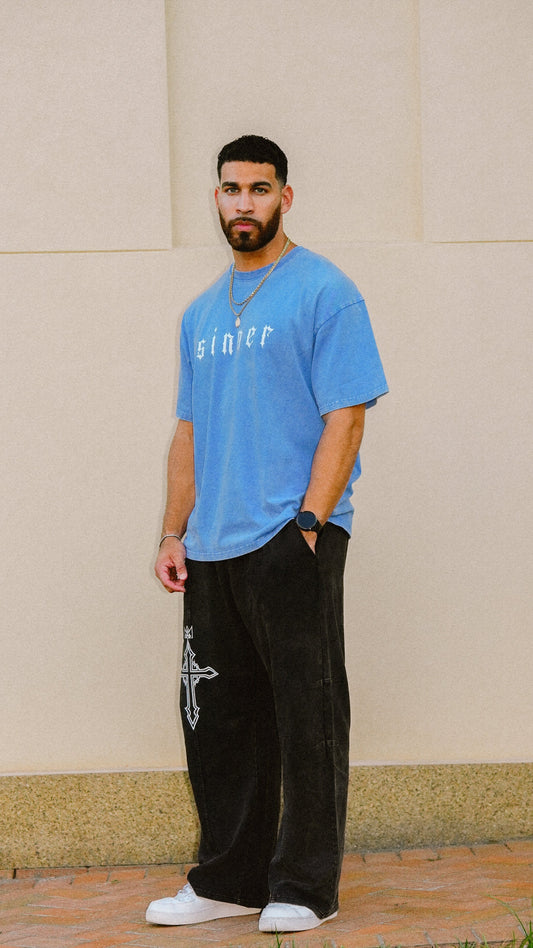 A man wearing a light blue t-shirt with 'sinner' printed on the chest, black wide-leg pants with a white cross graphic, and white sneakers, standing against a beige wall.