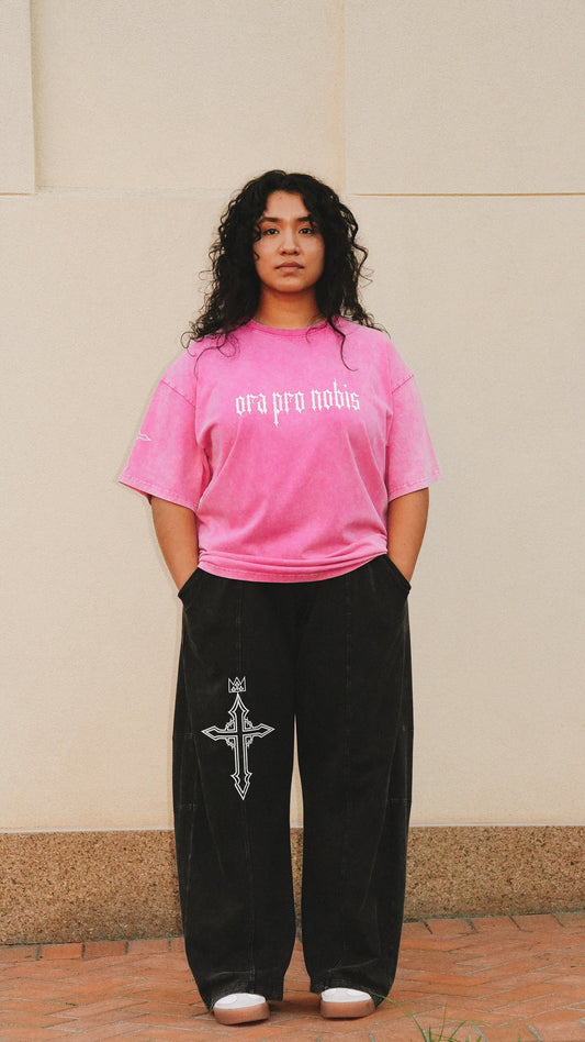 A person standing outdoors in front of a beige wall, wearing an oversized bright pink t-shirt with 'ora pro nobis' printed in white across the chest, paired with loose black sweatpants featuring a white cross graphic on the left leg, and white sneakers.